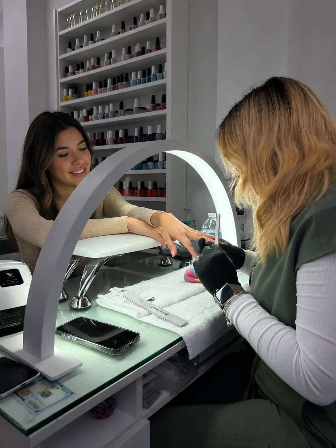 Manicure station with ring lamp: technician filing while the client checks her phone
