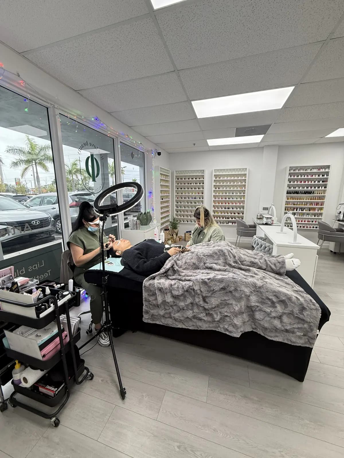 Interior of the Hialeah salon during a lash appointment: treatment bed with blanket, ring light, manicure stations and the polish wall behind